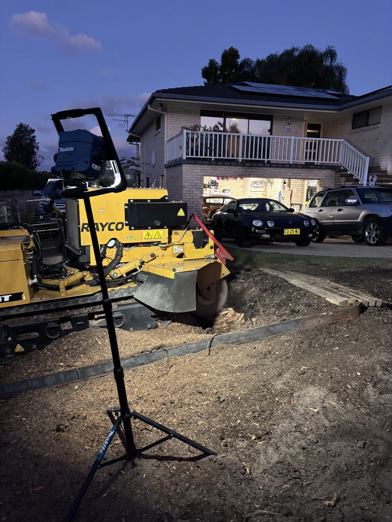 Stump grinder working beside a driveway at dusk under a portable work light