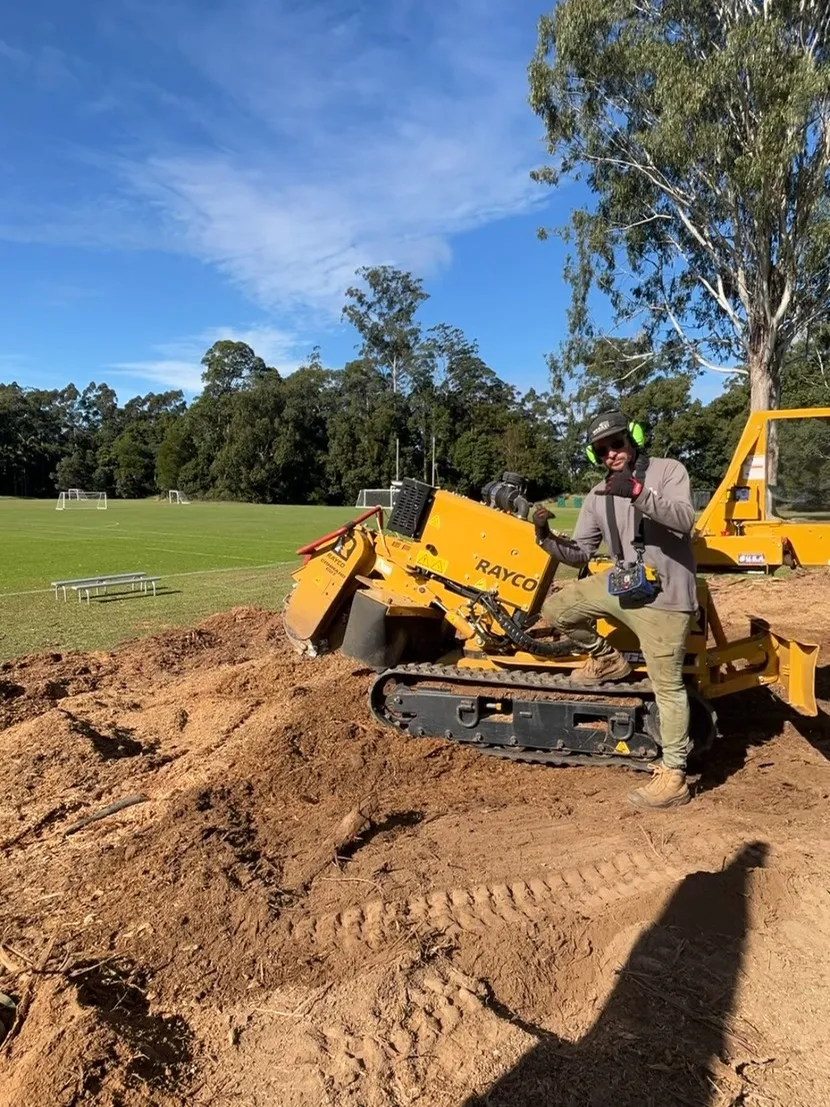 Stump Co. operator working the stump grinder on a cleared mound beside a sports field